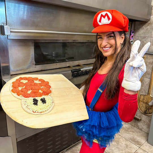 G dressed as Mario, holding a mushroom-shaped pizza on a peel beside a pizza oven