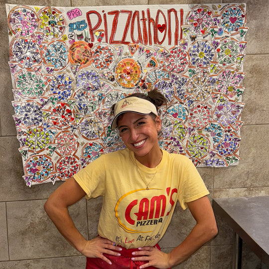 G in a Cam's Pizzeria tee and visor in front of the Pizzathon banner covered in pizza drawings